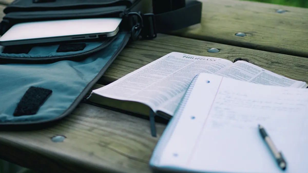 A student at a laptop pausing to think, notebook open beside it